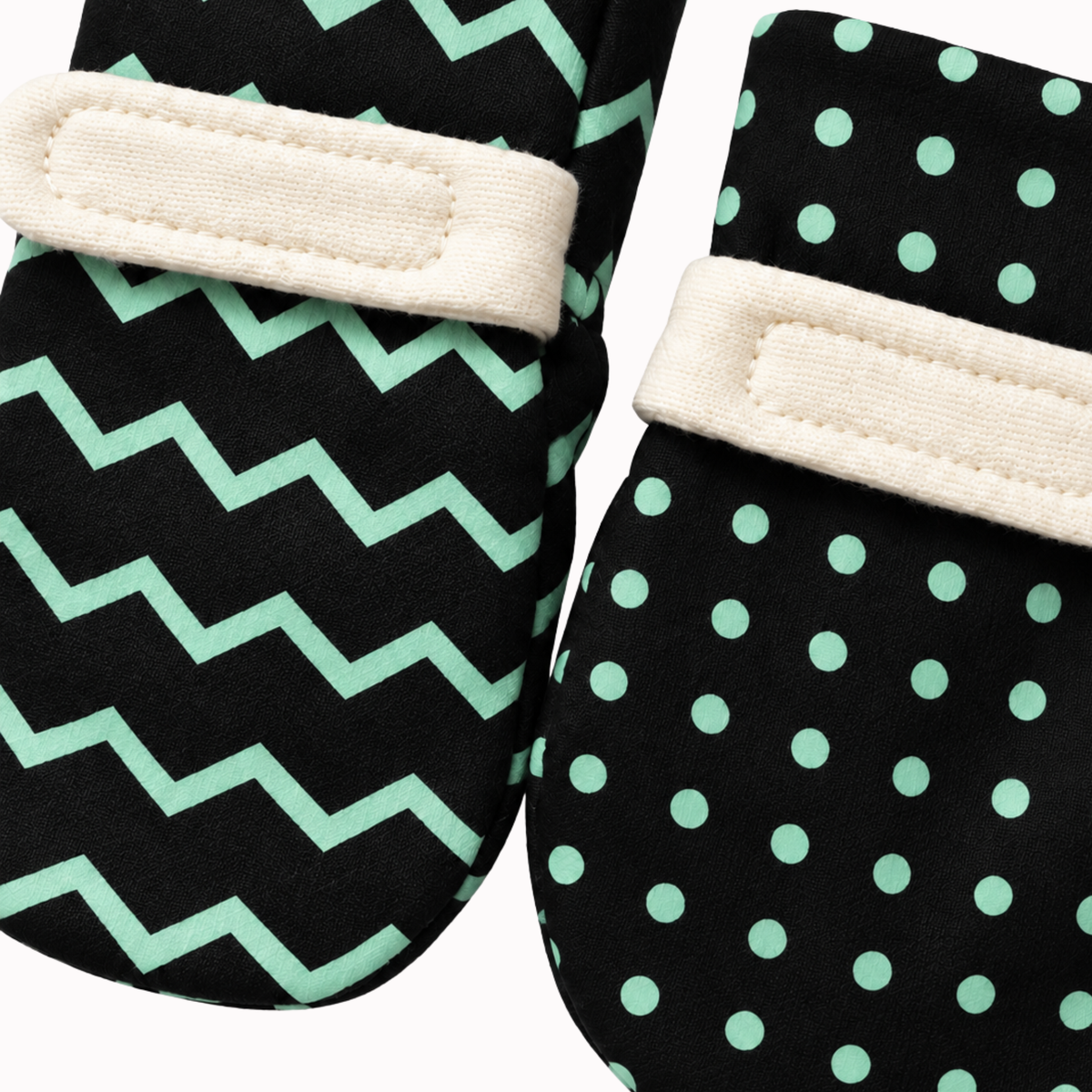 A pair of soft baby mittens with green visual patterns, featuring one mitten with a mint green chevron pattern and the other with a mint green polka dot pattern, both with cream fabric wrist straps.
