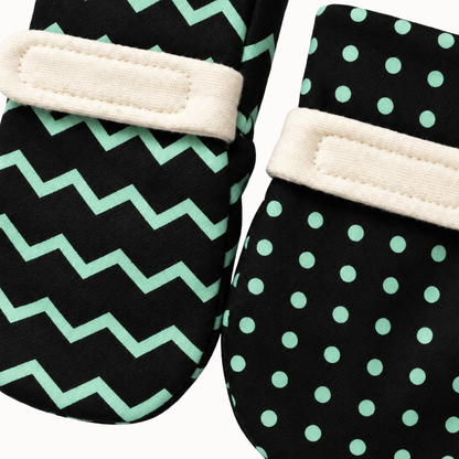 A pair of soft baby mittens with green visual patterns, featuring one mitten with a mint green chevron pattern and the other with a mint green polka dot pattern, both with cream fabric wrist straps.