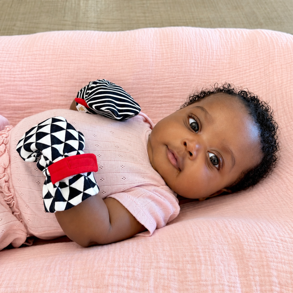 A baby lying on a pink textured blanket wearing a light pink outfit and black-and-white patterned soft baby mittens with red straps.