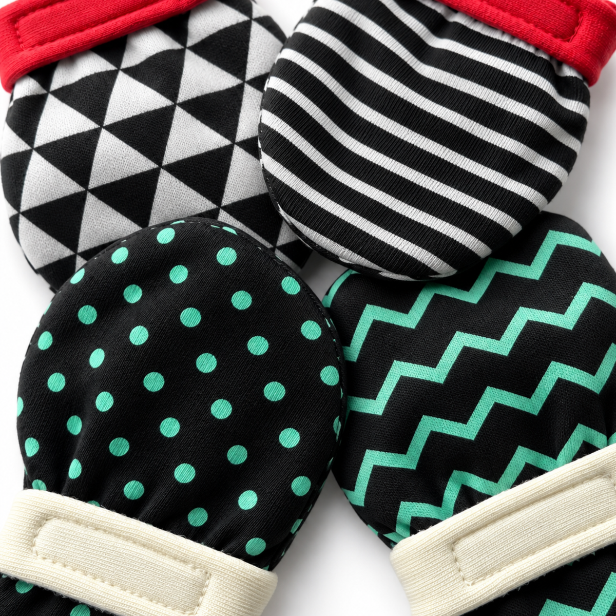 Four high-contrast baby mittens arranged in a flat-lay on a white background, each with a different pattern: black and white harlequin, black and white stripes, black with mint green polka dots, and black with mint green chevron. soft and 100% organic cotton. 