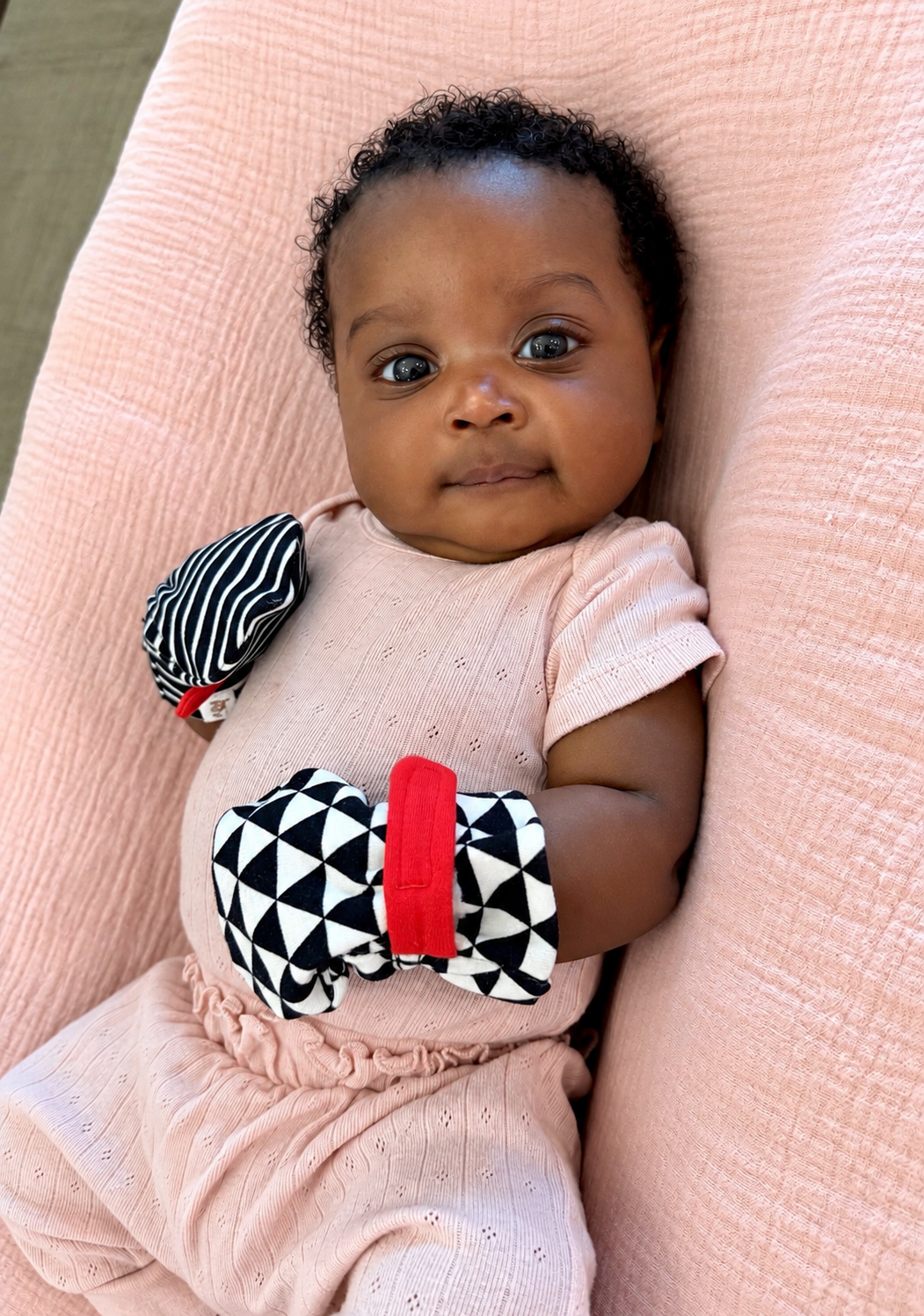 A baby wearing a light pink knit outfit and black-and-white patterned mittens with red accents, lying on a pink textured blanket. baby is wearing organic cotton mittens with a dual closure system. 