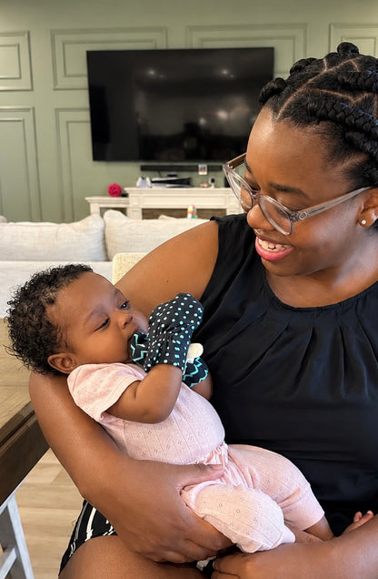 A mom holds her baby. The baby is wearing a high-contrast (black and teal polka-dot) mitten and looking at it. 